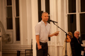 A man with a shaved head wearing black pants and a striped collared shirt reads from sheets of paper, speaking into a standing microphone. Two figures sitting at a table behind him turn to gaze towards him as he speaks.