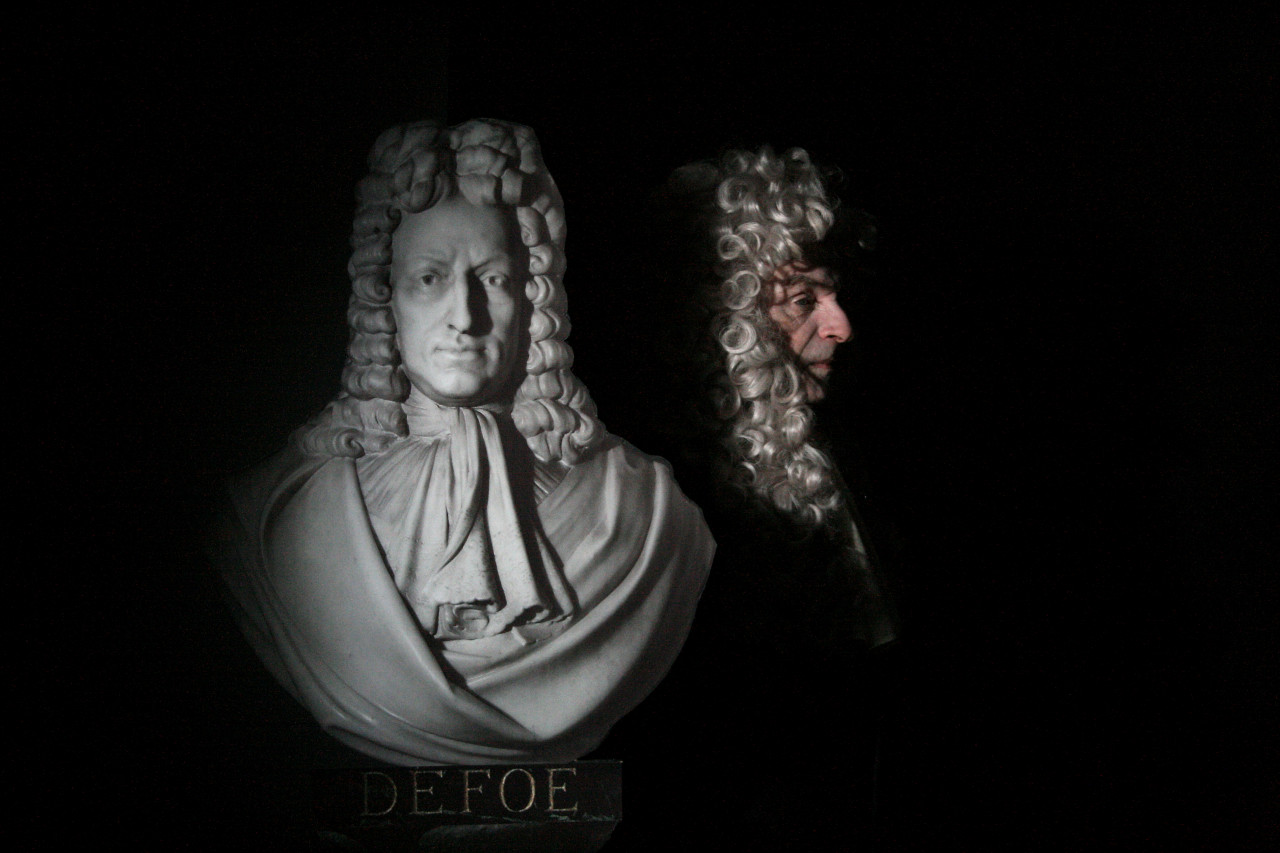 A white bust with a plaque reading "DEFOE" below it. Slightly offset and partially in shadow is a man seen in profile, dressed identically to the bust with a powdered wig.