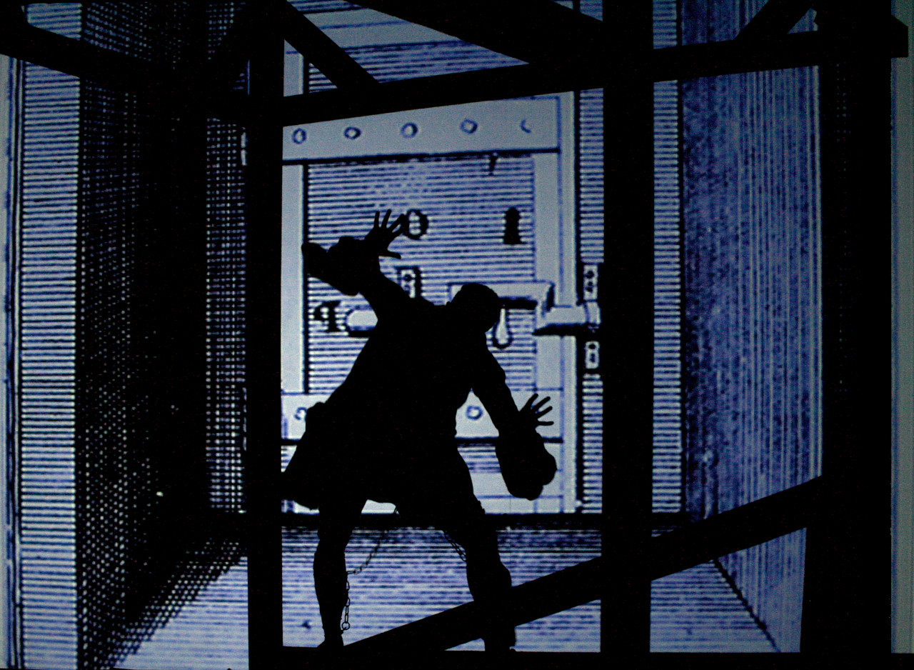 A animated cartoon in tones of black, blue, and grey depicting a silhouette of a prisoner with a chain on his leg and hands in the air, facing a large door with multiple locks.