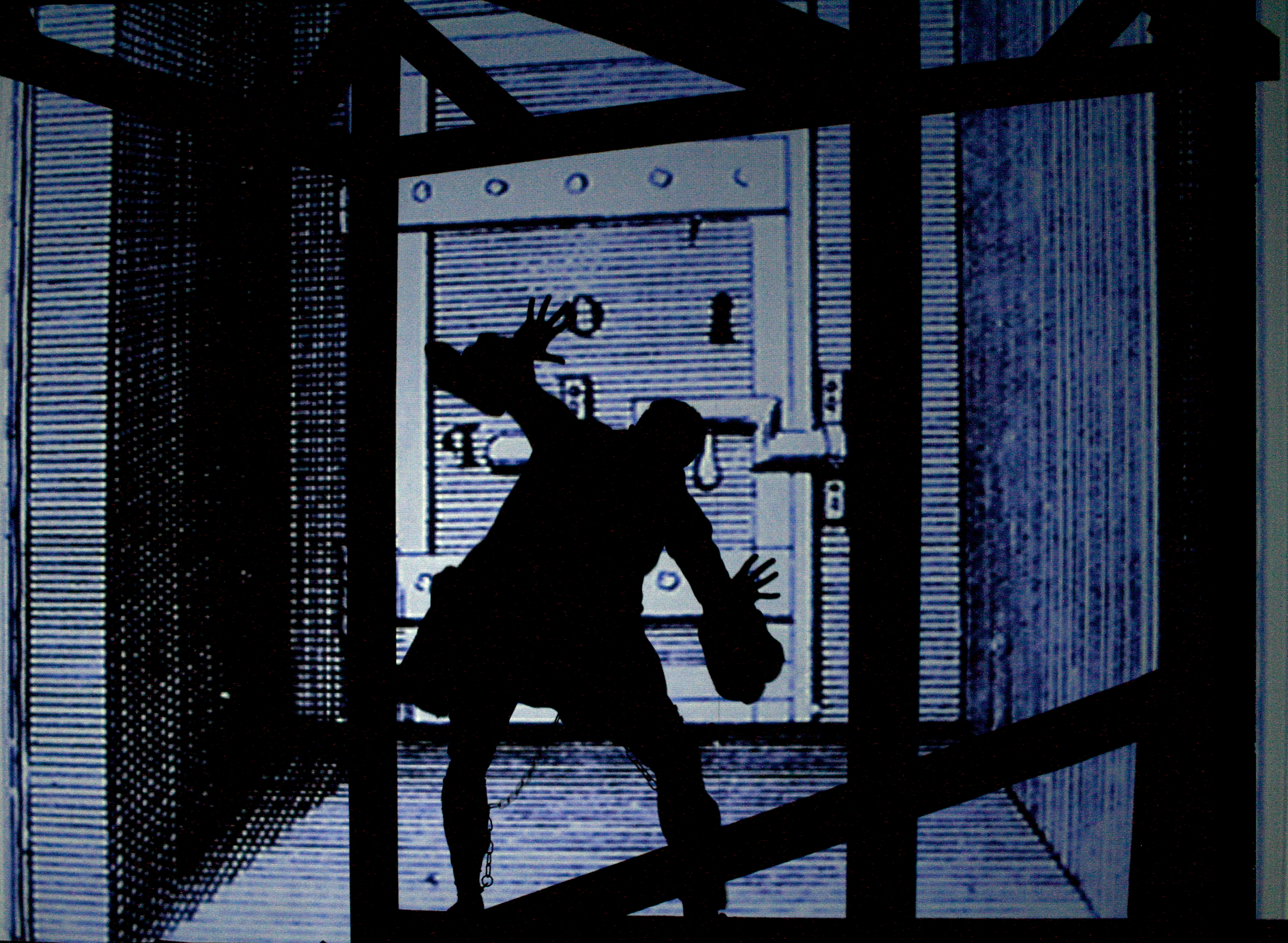 A animated cartoon in tones of black, blue, and grey depicting a silhouette of a prisoner with a chain on his leg and hands in the air, facing a large door with multiple locks.