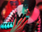A hand reaches toward the black-gloved hand of a person dressed in a black mask and jester-style costume, complete with a black and white pattern and a ruff around the neck.