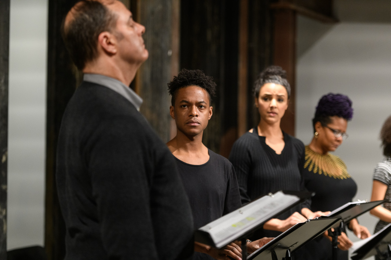 Five figures standing before a set of music stands. The figure closest to the viewer stands in profile and gazes ahead, while two figures directly behind them look towards him with furrowed brows.