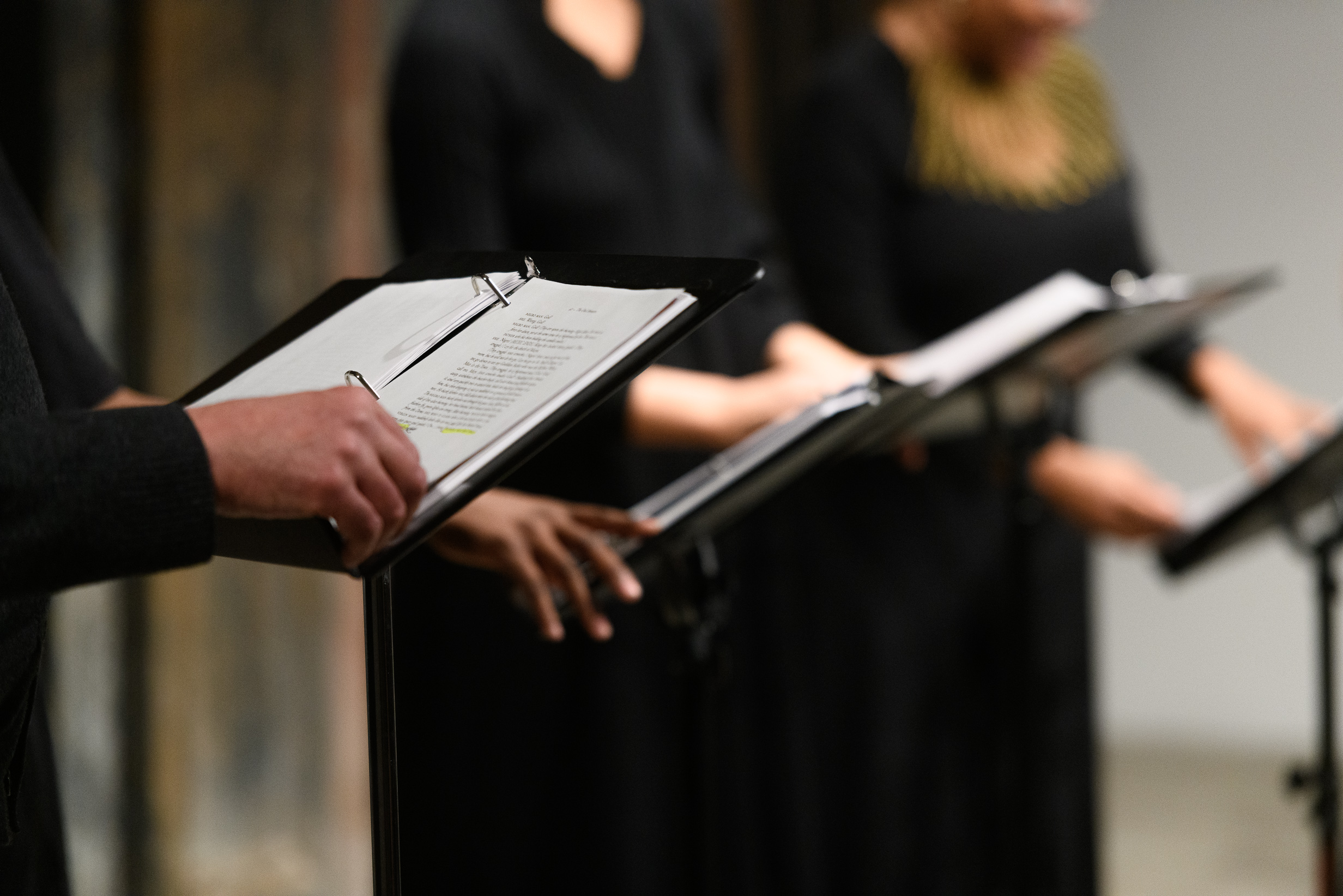 Close up shot of four music stands arranged side by side. The torsos and hands of three figures are pictured beside the stands. They leaf through the pages of scripts held in black binders, supported by the music stands.