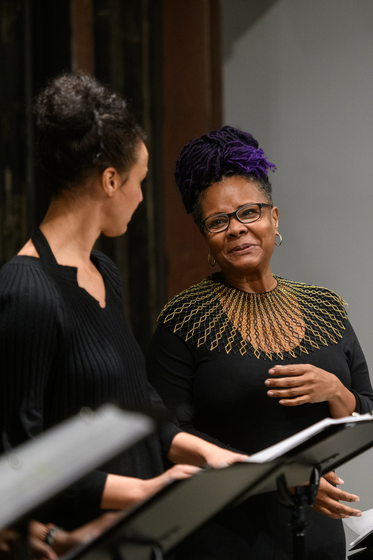 Close up of two figures standing before a set of music stands, their hands turning the pages of a script. They gaze at each other while the figure on the right speaks, their arm raised towards their chest in a small gesture. This figure wears glasses and an elaborate, gold and black collar piece.