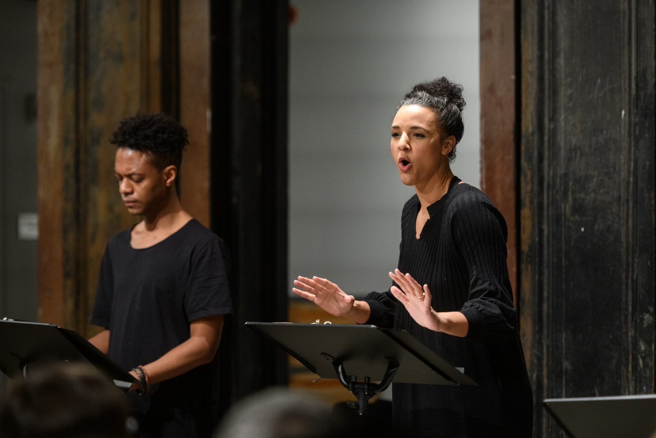 Two figures standing behind a set of music stands. The figure on the left gazes downwards, while the figure on the right looks up, speaking. Her hands are raised by her torso in a gesture. Both figures wear dark clothing.