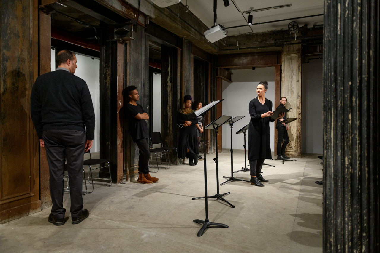 Six figures standing around a line of music stands in an interior space with concrete flooring and industrial columns. Some of the figures lean against the columns, their hands at their sides or casually crossed at their chests. Five of them stare at an isolated figure situated in front of the line of music stands, holding a black binder. She turns to gaze back at the others, illuminated by overhead lights as she speaks.