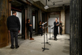 Six figures standing around a line of music stands in an interior space with concrete flooring and industrial columns. Some of the figures lean against the columns, their hands at their sides or casually crossed at their chests. Five of them stare at an isolated figure situated in front of the line of music stands, holding a black binder. She turns to gaze back at the others, illuminated by overhead lights as she speaks.