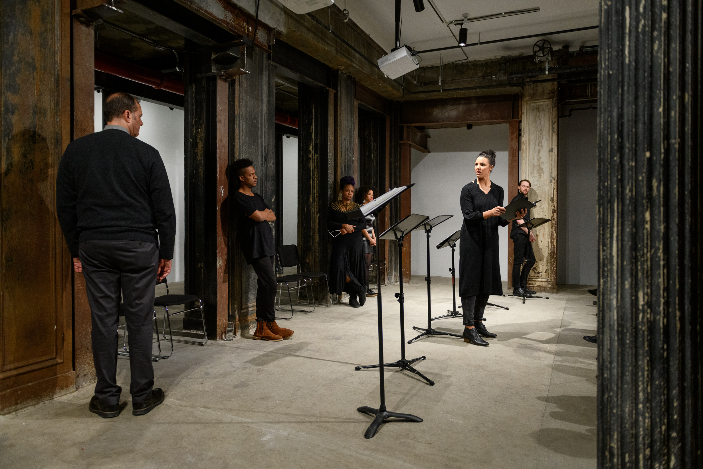 Six figures standing around a line of music stands in an interior space with concrete flooring and industrial columns. Some of the figures lean against the columns, their hands at their sides or casually crossed at their chests. Five of them stare at an isolated figure situated in front of the line of music stands, holding a black binder. She turns to gaze back at the others, illuminated by overhead lights as she speaks.