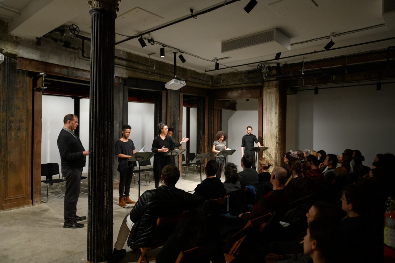 Six figures stand before a set of music stands in front of a seated audience in an interior space with industrial columns. They stand side by side in a horizontal line and are illuminated by overhead lights. A figure at the group