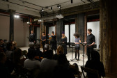 Six figures each stand before a music stand in front of a seated audience. They are arranged side by side in a horizontal line, illuminated by overhead lights. A figure at the group's center is speaking. Their outstretched hand gestures towards the crowd.