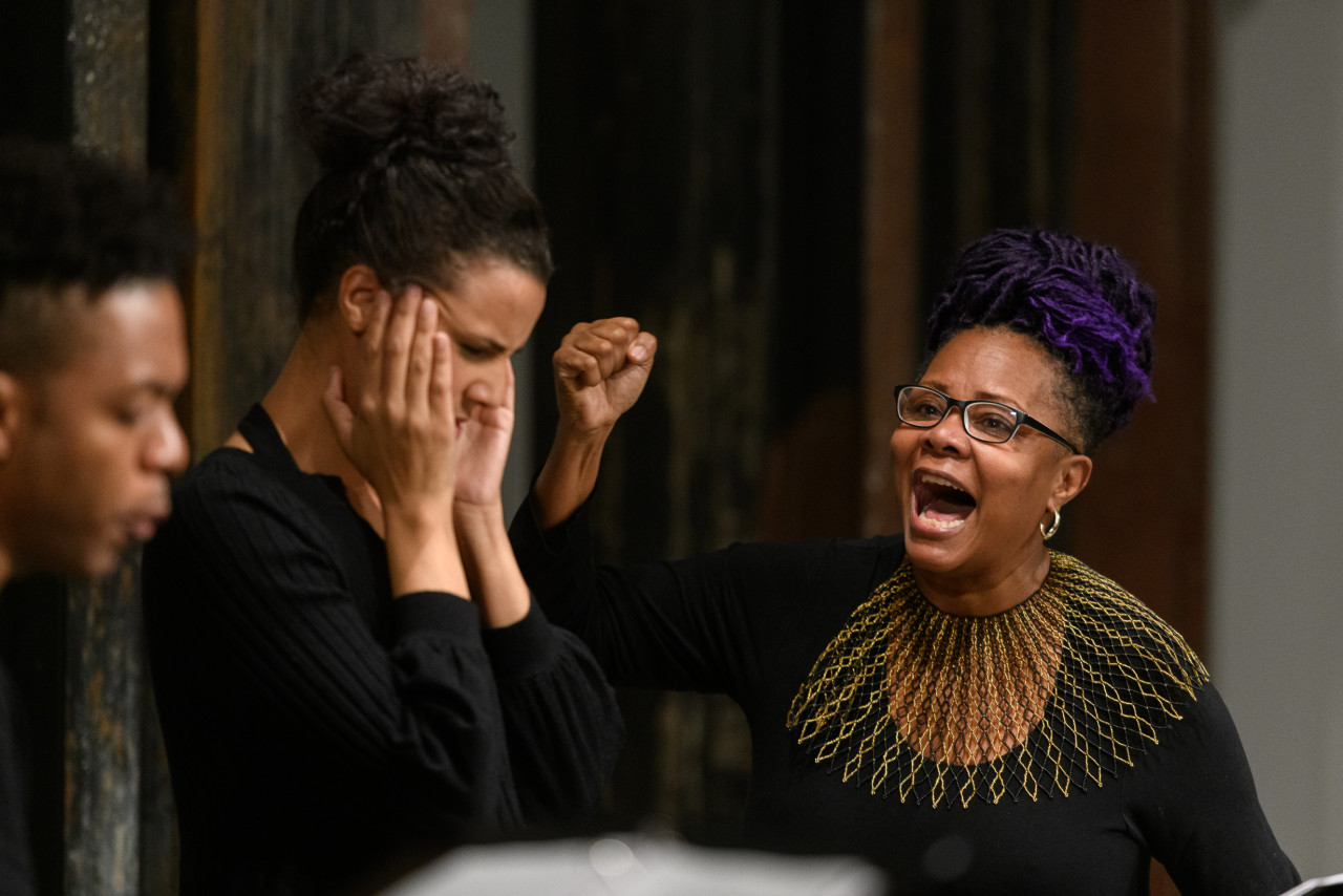 Close up of three figures standing side by side. The figure on the right raises a fist in the air and appears to shout towards the figures on their left. They wear glasses and an intricate, gold and black collar piece. The figure directly to their left gazes downwards, pressing their fingers to their temples.