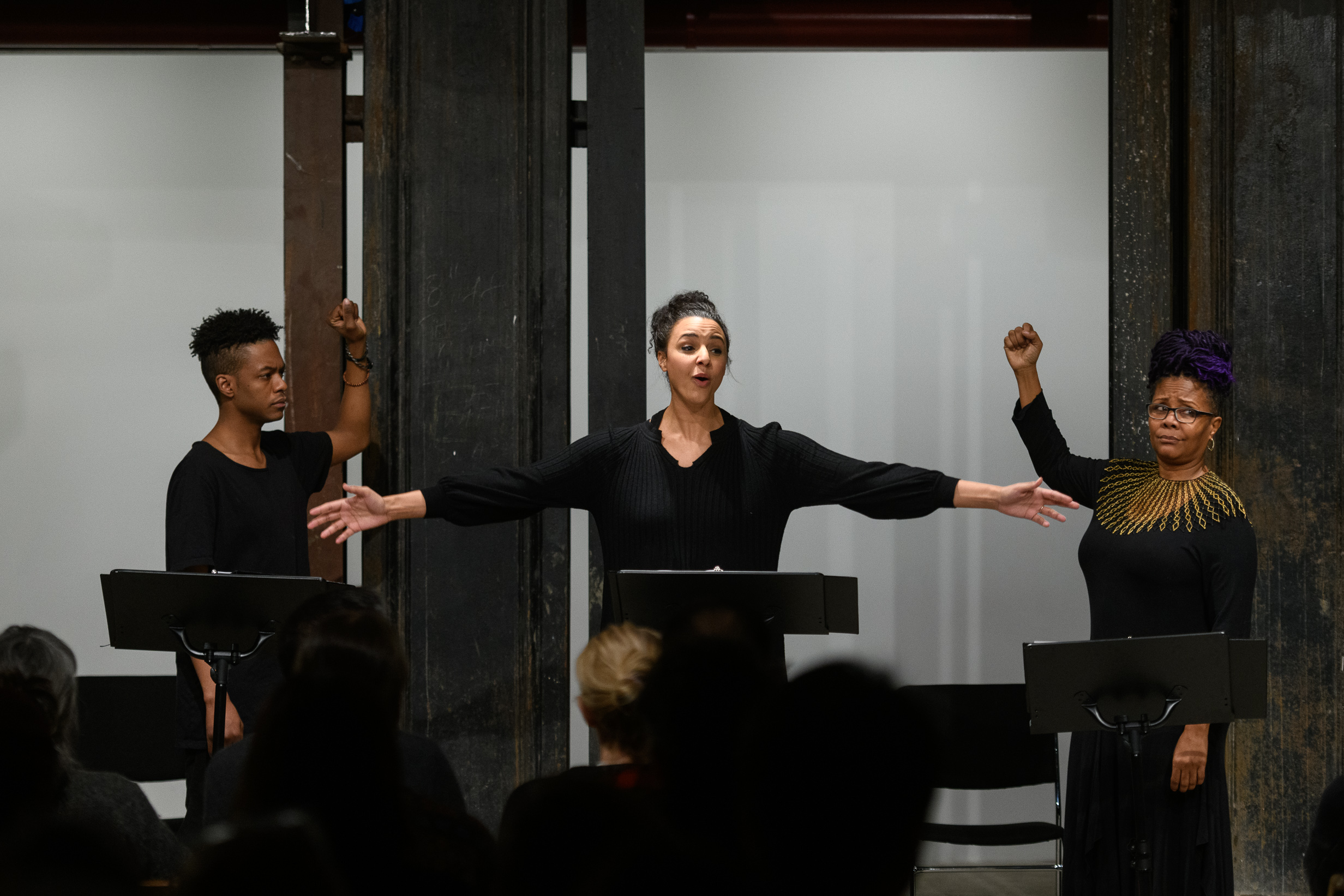 Three figures standing by a set of music stands in front of a seated audience. The center figure faces the audience, her arms stretched wide. The figures on either side of her appear to mime grasping something above themselves—such as a bar or handle—with one hand. One's expression is solemn, the other's incredulous.