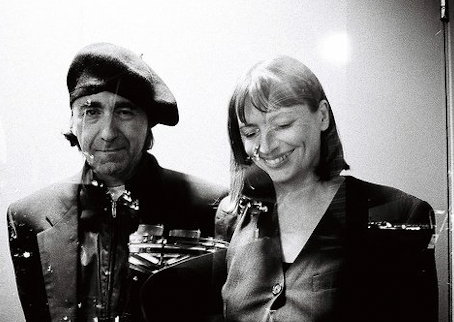 Black-and-white photograph that has been double exposed. The dominant image shows a portrait of two smiling figures, standing side-by-side. Imposed on top of that is a faint image of a building and lights.