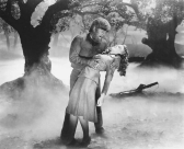 A black and white image captures the dark silhouette of trees shrouded with thick fog. In the center stands a man with wolf-like features, grimacing over a woman who is seemingly unconscious in his arms. Her body is arced back, supported by the wolf man's arm under her shoulders and around her waist.