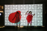 Two women in dresses and matching shoes are splayed up against a screen with their arms raised. There is an image projected over them, with a white background and black geometric pattern, and two splotches of red over their bodies.