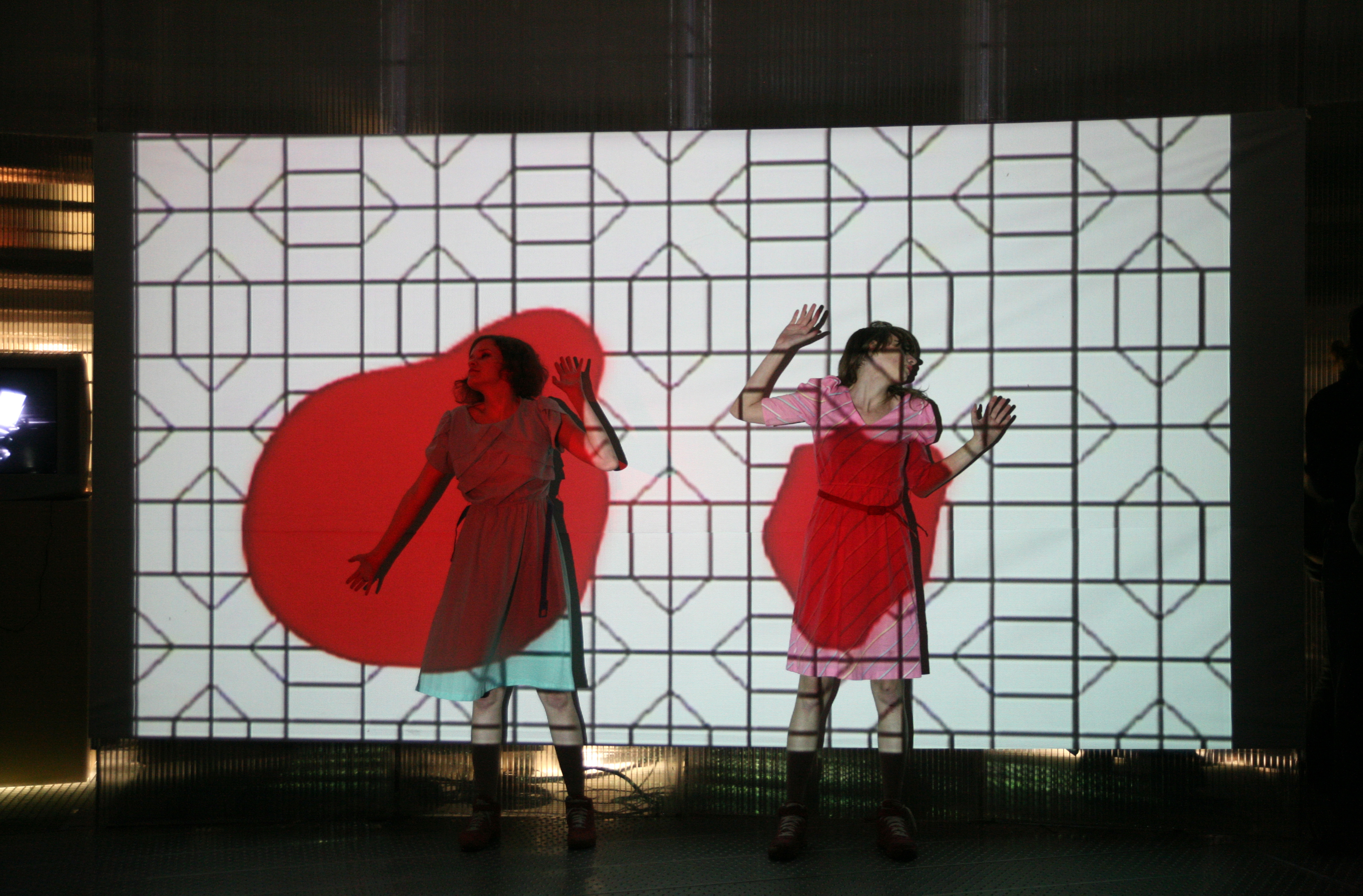 Two women in dresses and matching shoes are splayed up against a screen with their arms raised. There is an image projected over them, with a white background and black geometric pattern, and two splotches of red over their bodies.