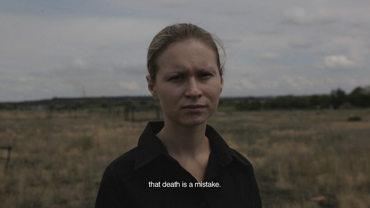 A film still of a woman, captured from the shoulders up, standing in an outdoor field. The white subtitle reads "that death is a mistake."