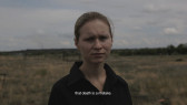 A film still of a woman, captured from the shoulders up, standing in an outdoor field. The white subtitle reads 