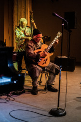 A figure plays the guitar sitting down, while another figure stands behind him playing the bass.
