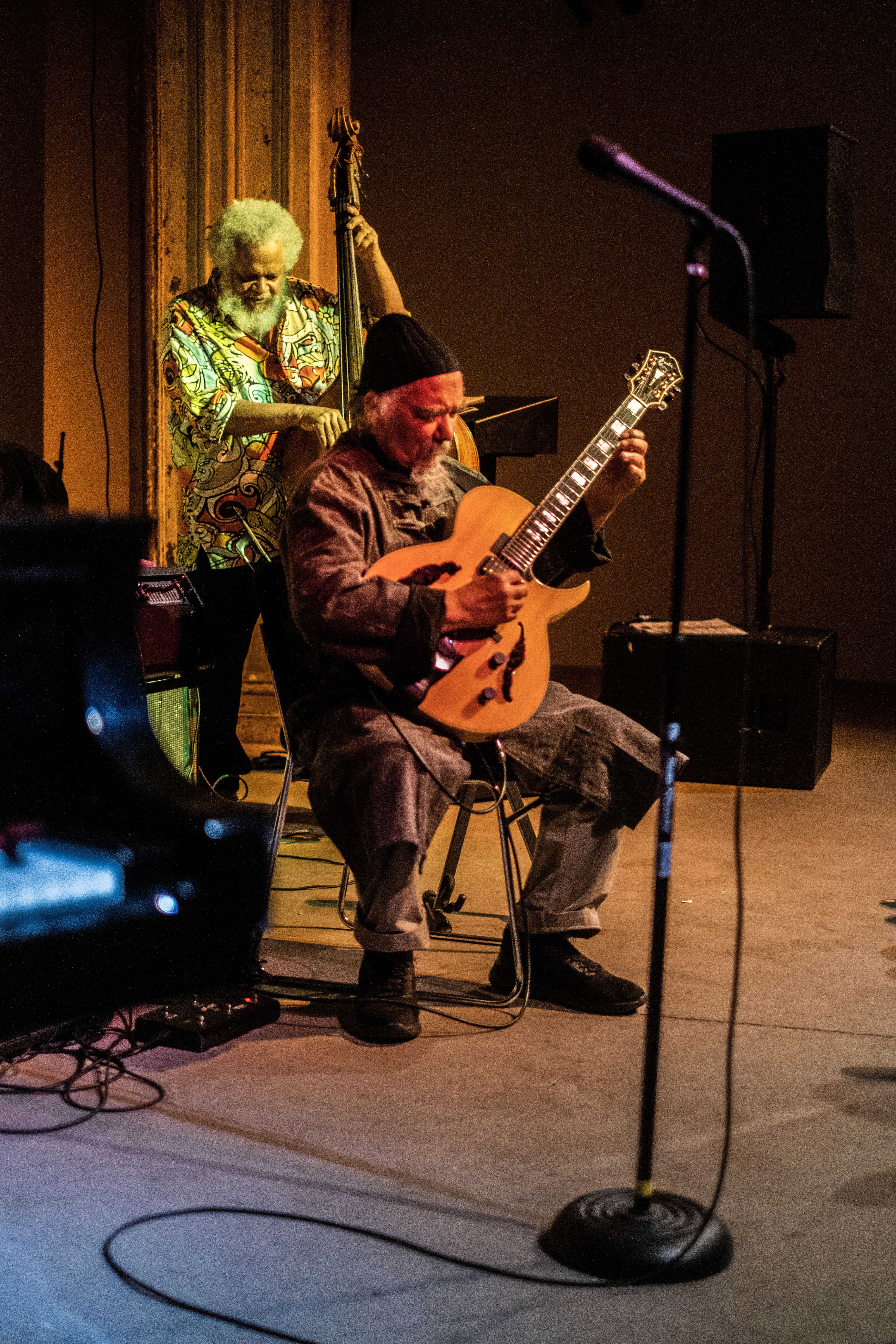 A figure plays the guitar sitting down, while another figure stands behind him playing the bass.