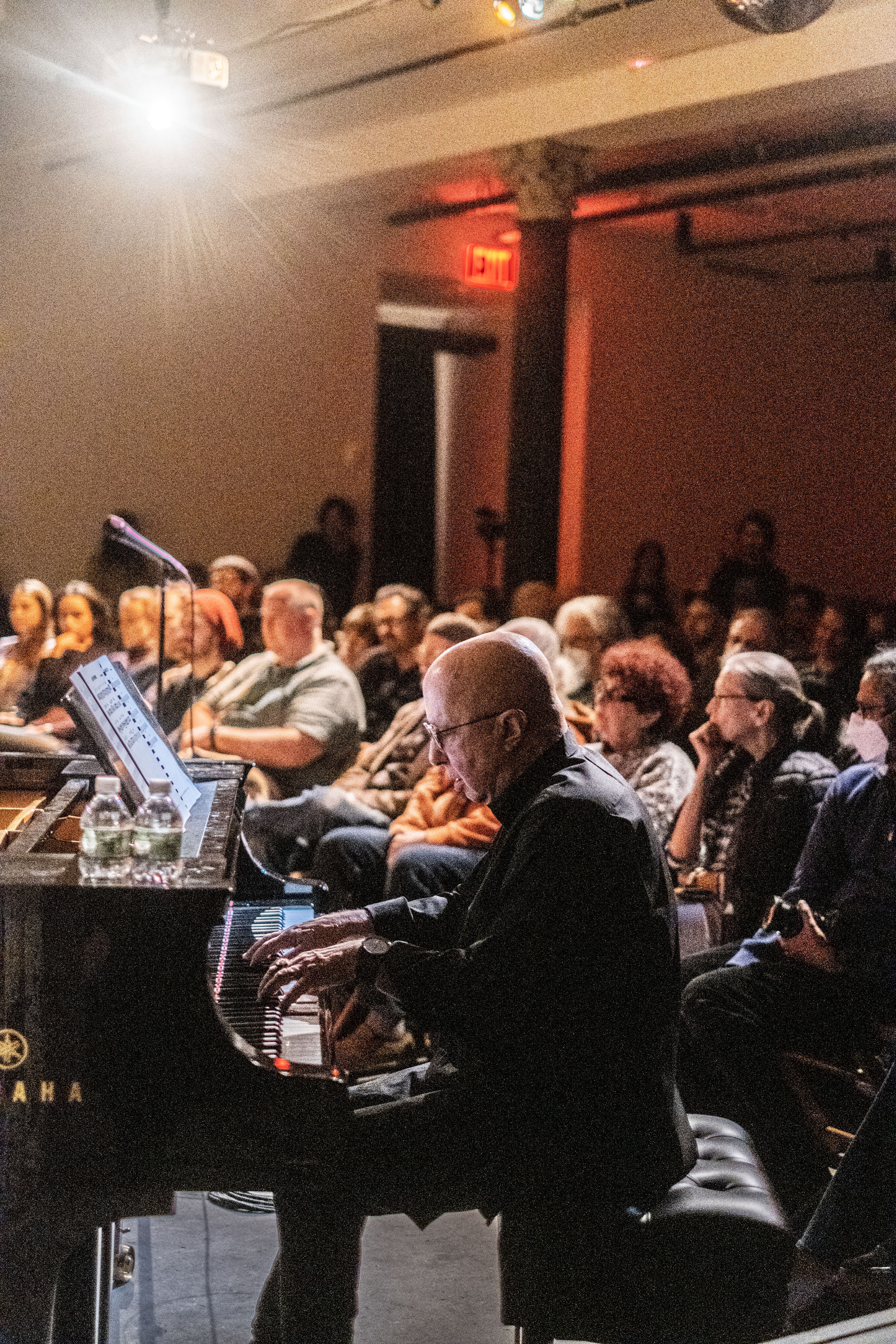 A figure plays the piano to a seated crowd behind him.