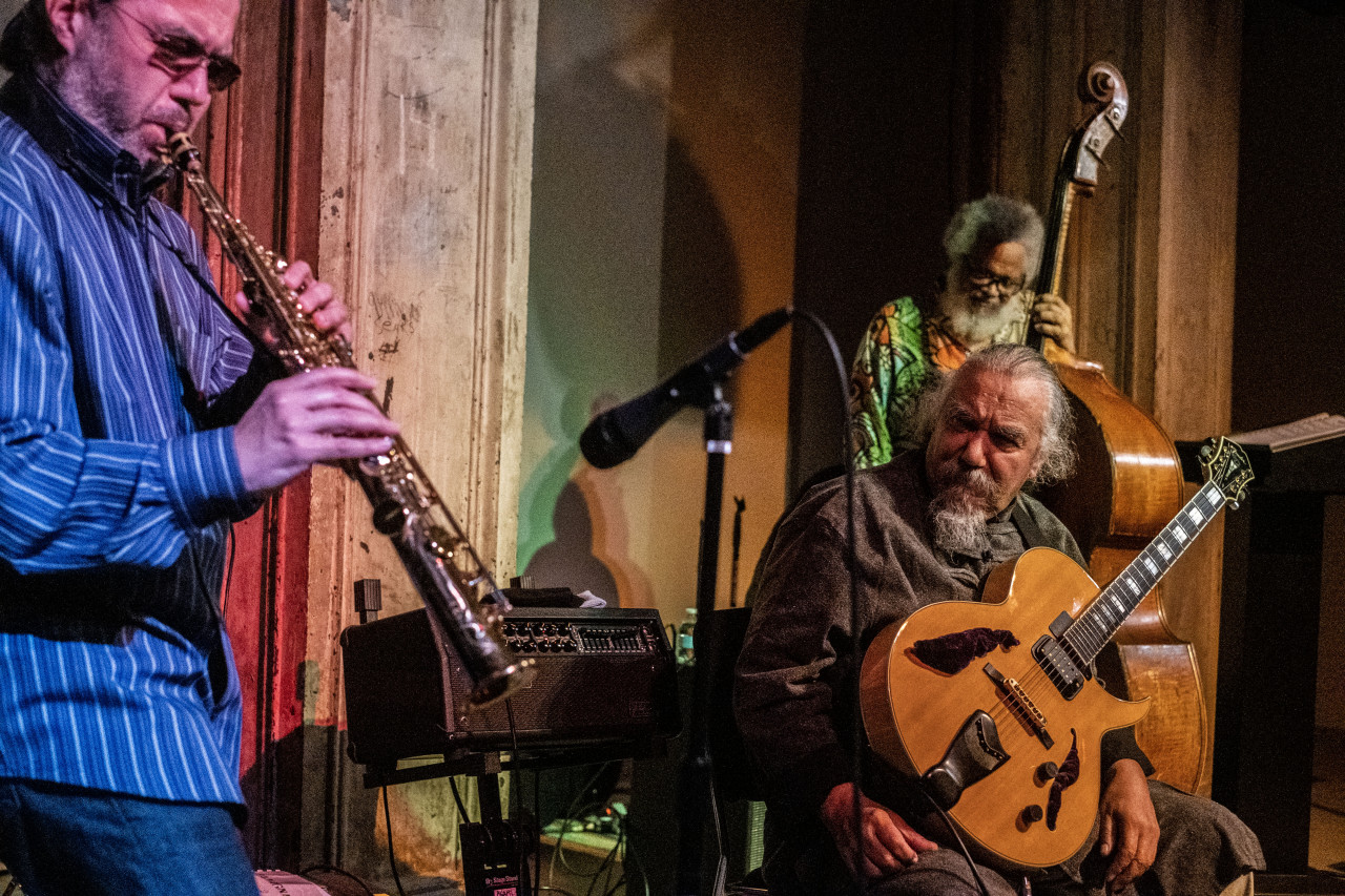 A figure to the left plays the saxophone while another figure sitting in the middle of the frame watches with a guitar in his lap, another figure in the back right plays the bass.