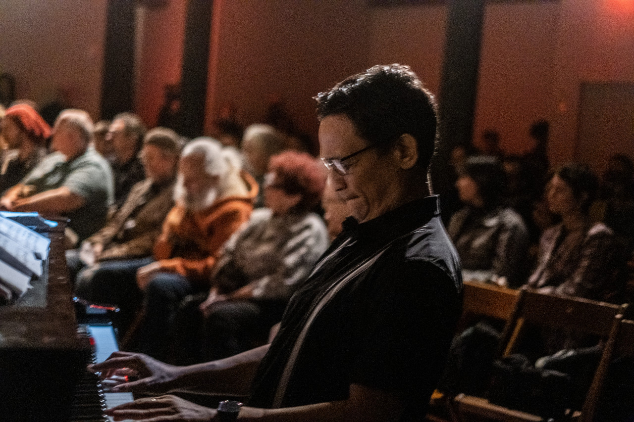 A figure plays the piano in front of a crowd of seated people behind him.