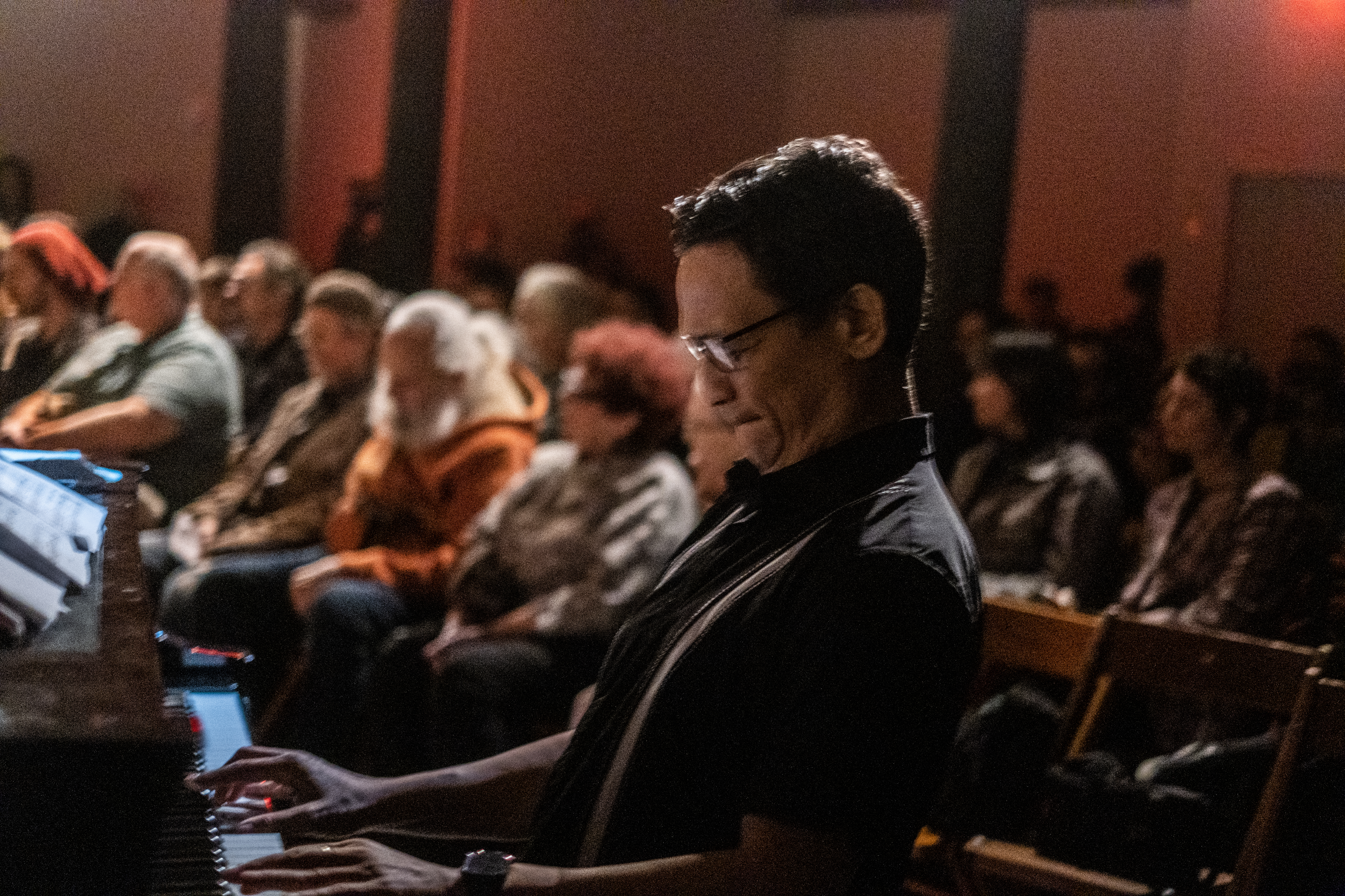 A figure plays the piano in front of a crowd of seated people behind him.