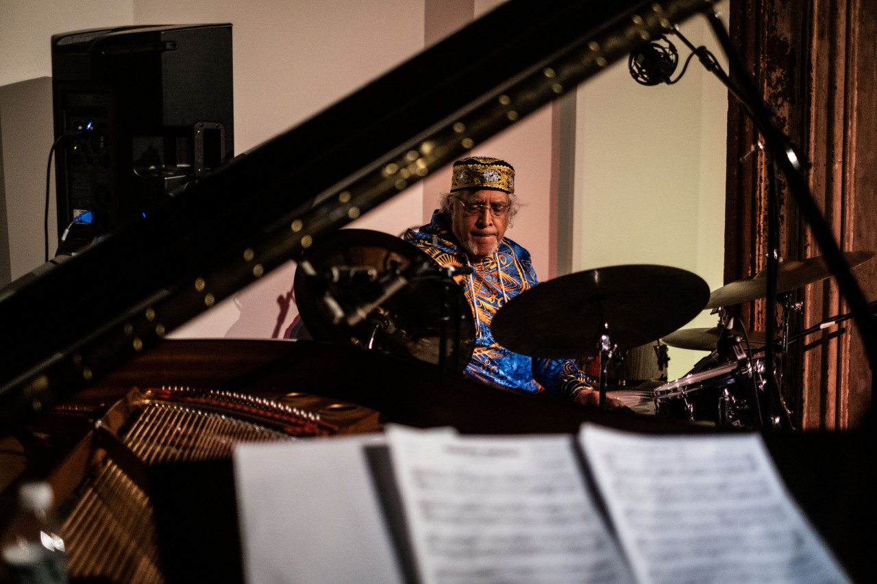 A figure plays the drums in the background with an out of focus piano in the foreground.