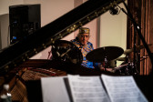 A figure plays the drums in the background with an out of focus piano in the foreground.