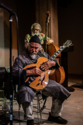 A figure plays the guitar sitting down, while another figure stands behind him playing the bass.
