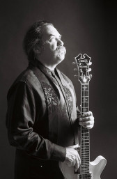 A black and white image of a figure in profile with long, wispy hair and a mustache holding a guitar.