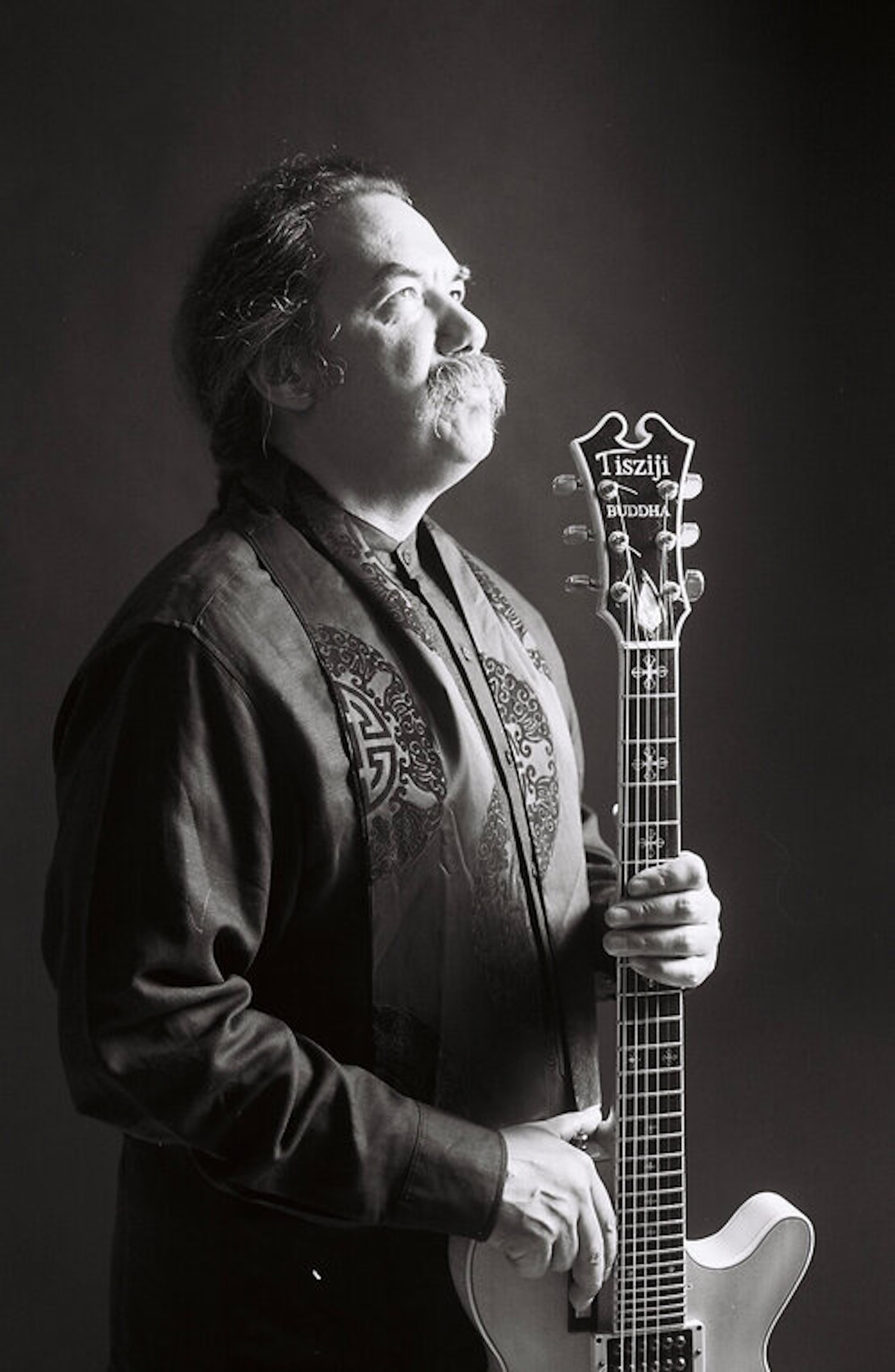 A black and white image of a figure in profile with long, wispy hair and a mustache holding a guitar.