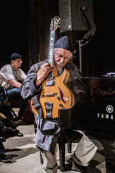 A figure sits on a piano bench, holding a guitar vertically against the figure's body.