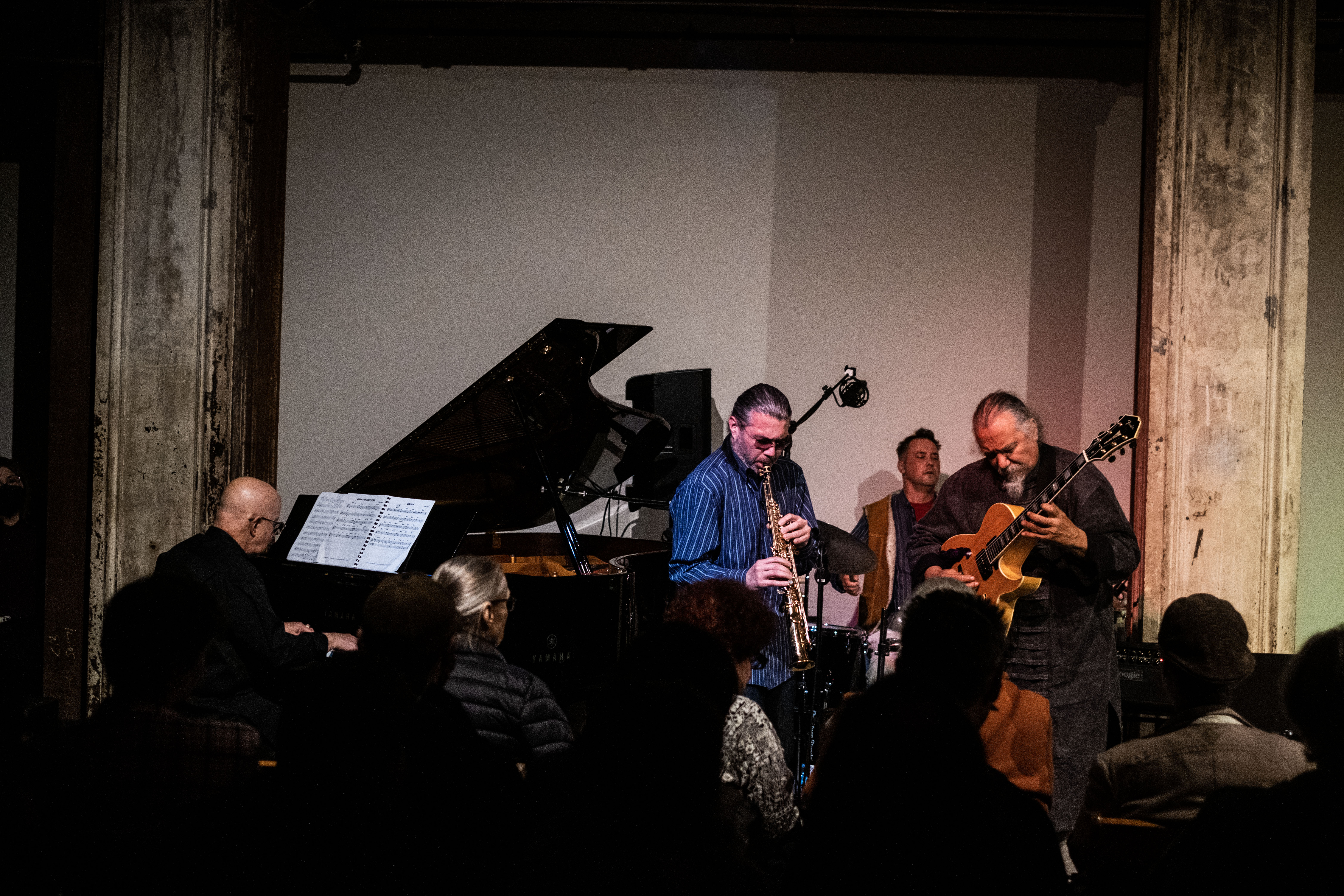 A band plays on stage in front of a seated crowd. From left to right, a figure is sitting and playing the piano, another figure stands and plays the saxophone, another figure in the back plays the drums, and the fourth figure stands and plays the guitar.