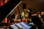 A band plays on stage, with a figure playing the piano in the foreground of the image, while another plays congas behind him. A third figure plays the tuba in the background.