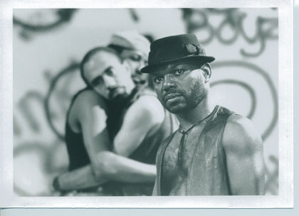 Two men embrace in the background, while a man wearing a black top hat stands in the foreground gazing at the camera.