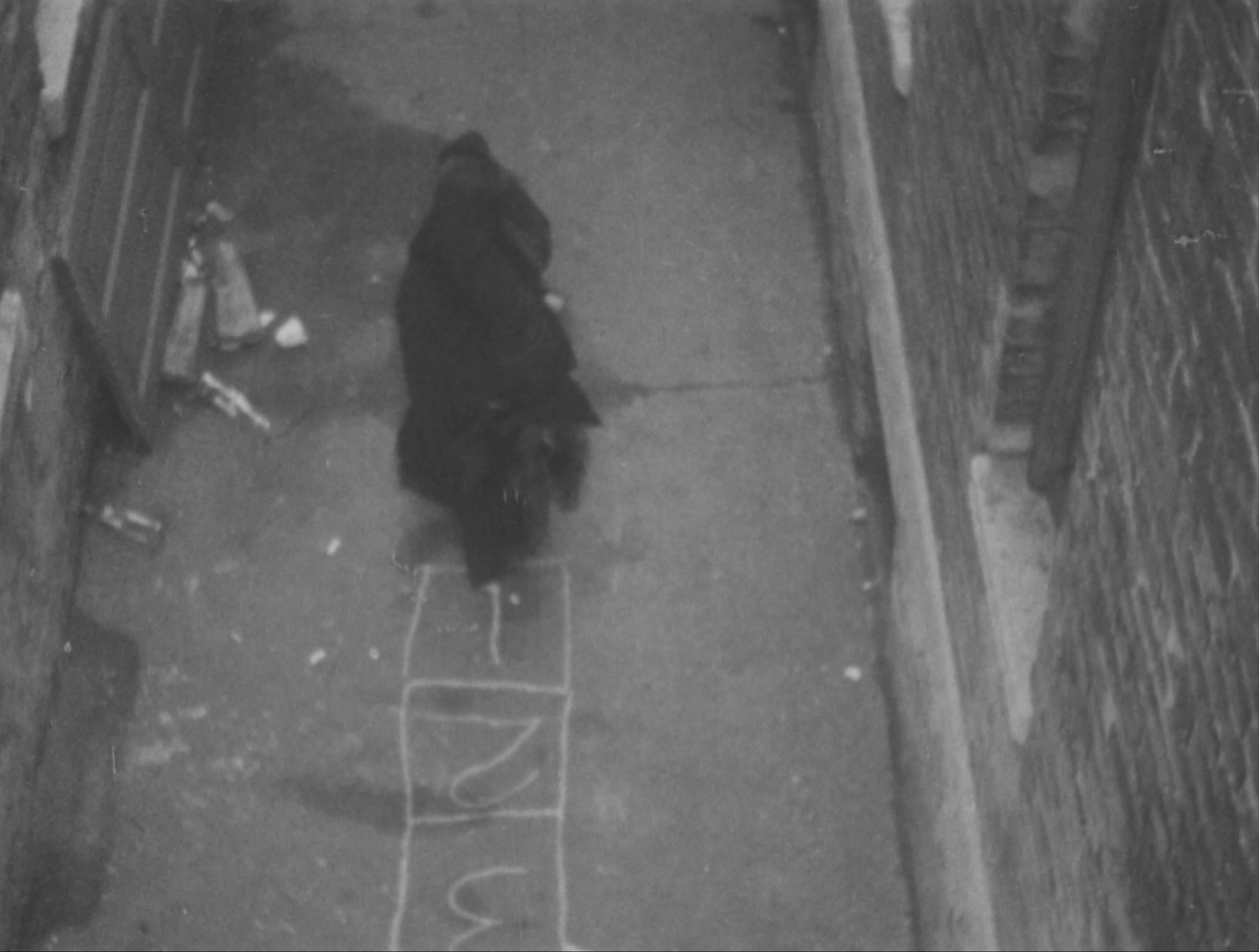 Black-and-white image of a figure in a long black coat bending down and drawing a hop scotch pattern in an alleyway using white chalk.