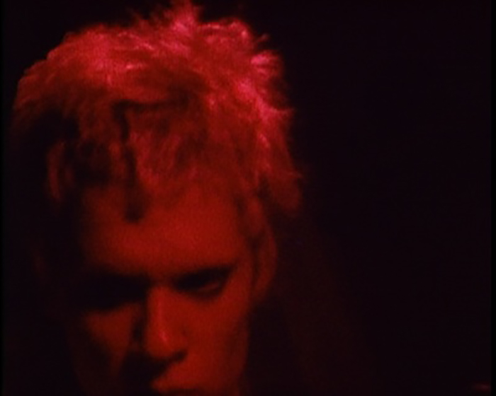 Close-up of the face of a figure with short, light hair. The figure looks downwards. They are illuminated by a dim red light.