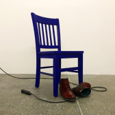 A pair of leather boots and a power strip rest underneath a blue, wooden chair on a concrete floor.