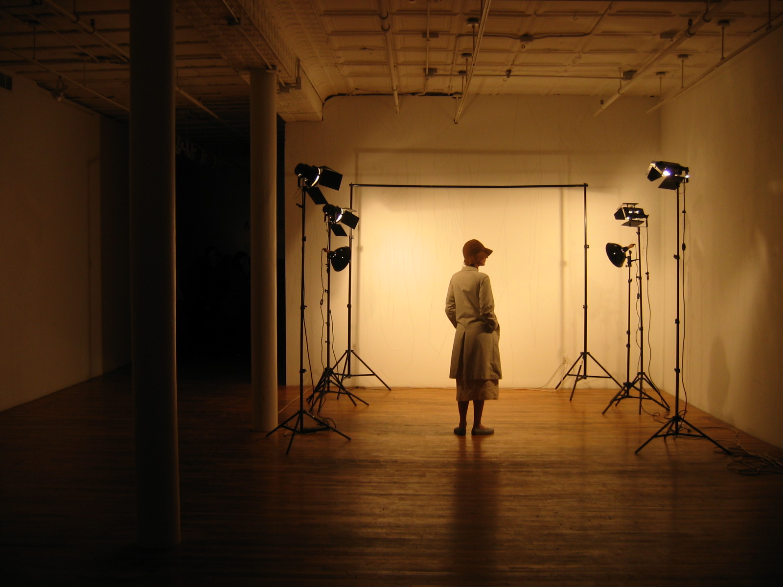 A darkened gallery room with several standing lights pointing to illuminate a corner. In the corner, a figure stands, wearing a coat and a hat with her hands in her pockets, facing away from the camera. She is framed by an empty metal stand.