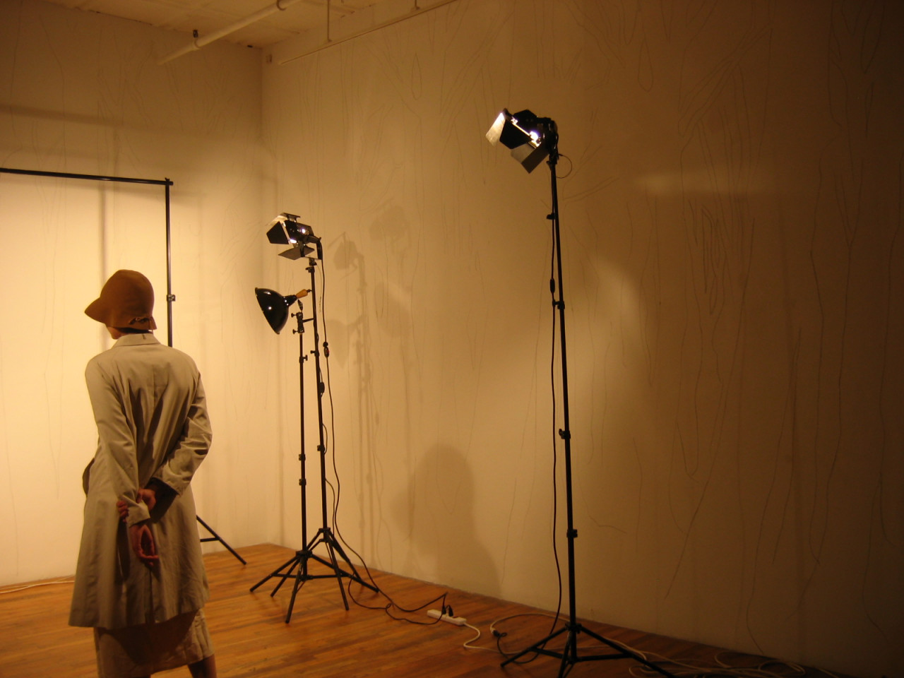 Three standing lights point to illuminate a white wall. Standing beside them facing the wall is a figure wearing a coat and a hat with her hands lightly clasped behind her back. Barely visible on the wall beside her are stenciled outlines of trees.