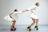 Two women in front of a white wall. They are dressed in matching white jackets and short white skirts, and each wear a set of colorful rollerskates. One woman is on her toes with her arms extended behind her, reaching for and pulling the second woman who is leaning forward to grab her hands.