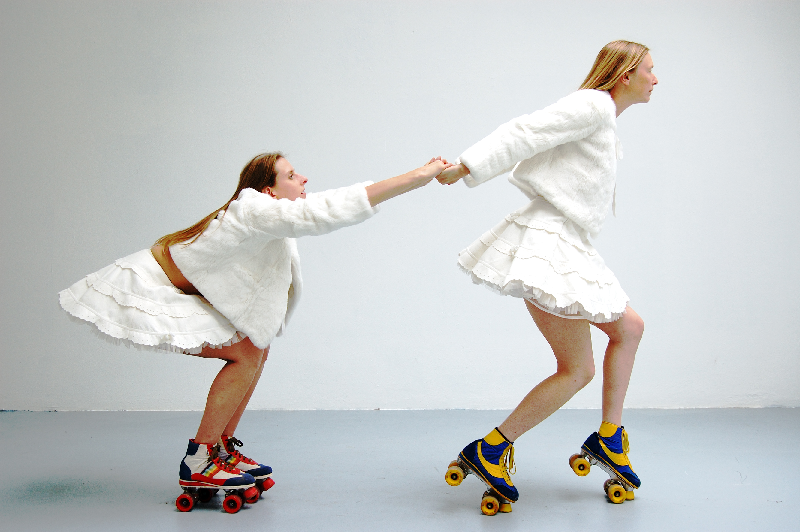 Two women in front of a white wall. They are dressed in matching white jackets and short white skirts, and each wear a set of colorful rollerskates. One woman is on her toes with her arms extended behind her, reaching for and pulling the second woman who is leaning forward to grab her hands.