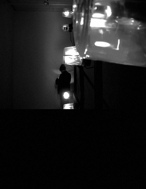 A black and white image of a figure wearing all black, with a black hat, facing to the wall and slightly illuminated by a light in their hands. In the foreground, there is a partially visible metal grid with several plastic-covered white lights.