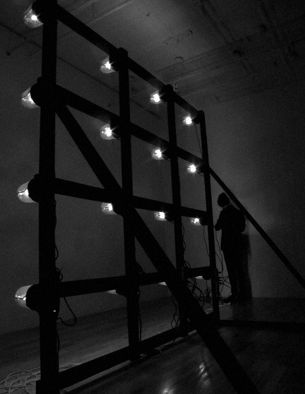 A black and white image of a three by four metal grid with plastic-covered white lights on each intersection. To the right of the grid is a figure wearing all black, with a black hat, facing to the wall.