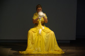 A woman is seated wearing a long, yellow dress with a full skirt that has extra fabric at the bottom. She is holding up soapsuds, which are dripping down onto the front of her skirt.