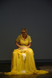 A woman is seated wearing a long, yellow dress with a full skirt that has extra fabric at the bottom. She is washing the front of her dress with soap suds, some of which are dripping down the front of the skirt.