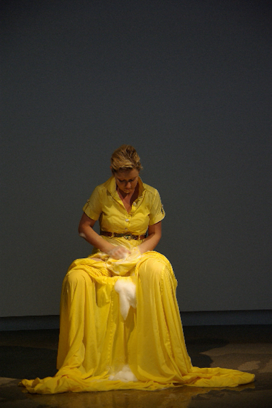 A woman is seated wearing a long, yellow dress with a full skirt that has extra fabric at the bottom. She is washing the front of her dress with soap suds, some of which are dripping down the front of the skirt.