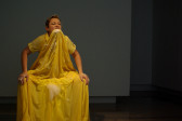 A woman is seated wearing a long, yellow dress with a full skirt. She has the front of the skirt held in her teeth, with her hands on her knees. There are some soap suds on her hands, near her mouth, and on the front of the skirt.